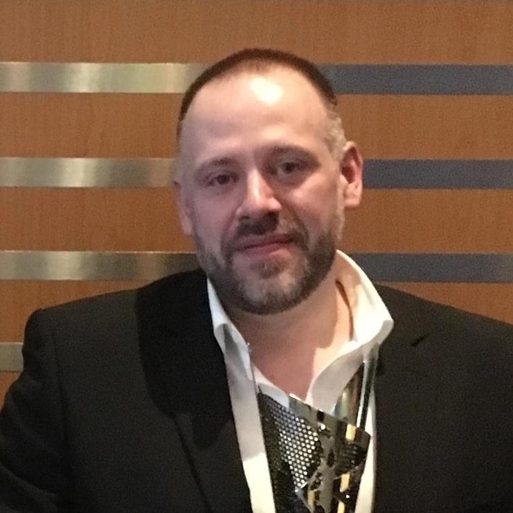 Mann in schwarzem Anzug mit Krawatte und Medienauszeichnung vor einem houten Hintergrund.