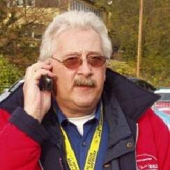 Mann mit Brille und grauen Haaren spricht am Handy und trägt eine rote Jacke sowie eine gelbe Umhängeleine.