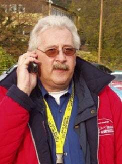 Mann mit Brille und grauen Haaren spricht am Handy und trägt eine rote Jacke sowie eine gelbe Umhängeleine.