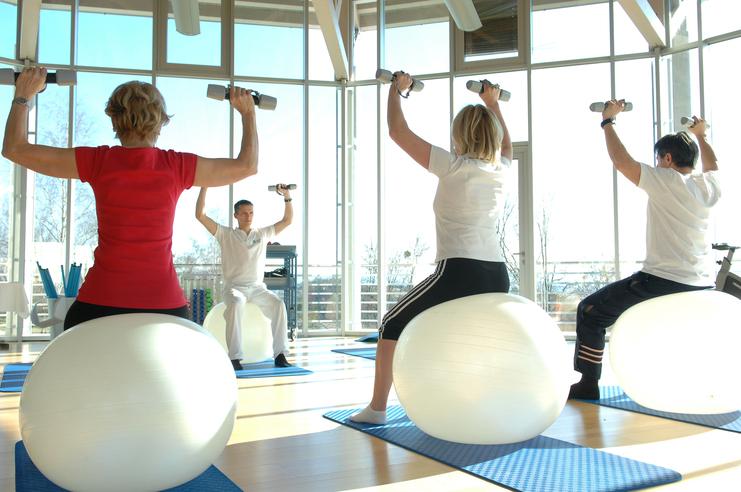 Gruppe von vier Personen trainiert auf Gymnastikbällen mit Hanteln in einem hellen Fitnessraum.