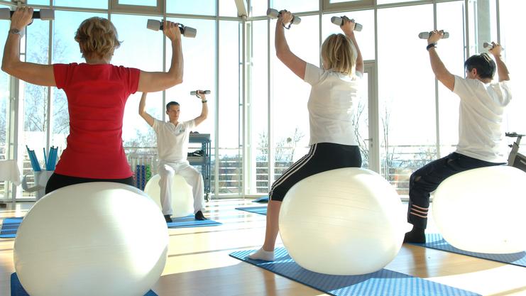 Gruppe von vier Personen trainiert auf Gymnastikb&auml;llen mit Hanteln in einem hellen Fitnessraum.