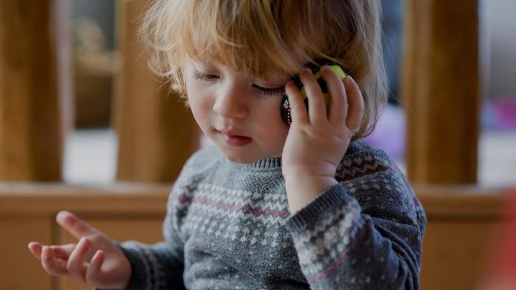 Kleines Kind mit lockigem Haar hält ein Spielzeugtelefon an das Ohr und schaut nachdenklich. Hintergründe sind unscharf.