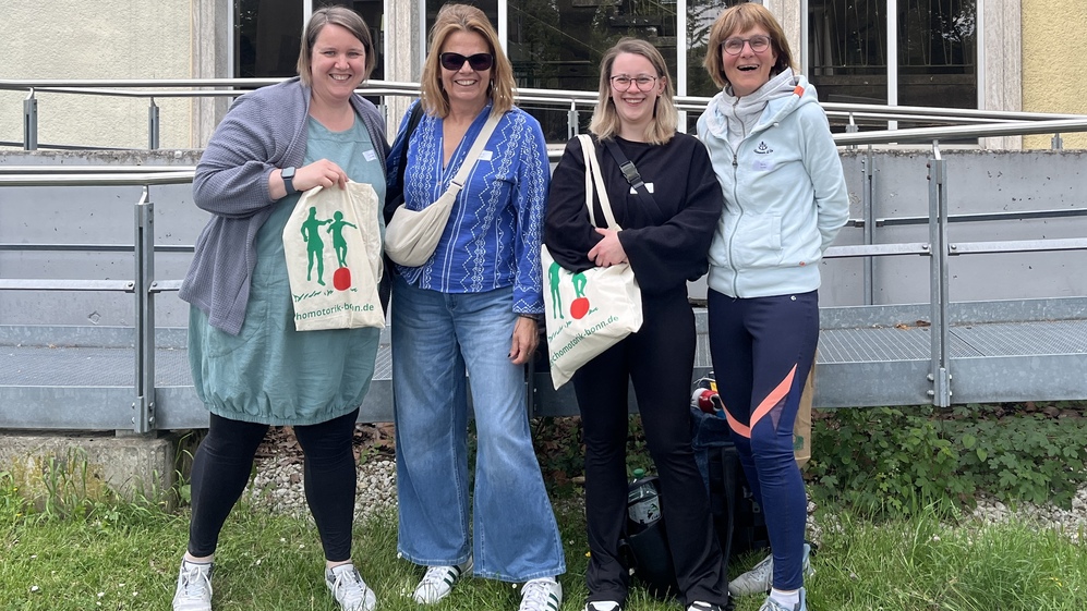 Vier Frauen stehen lächelnd vor einem Gebäude mit großen Fenstern, jede hält eine Tasche mit einem bunten Motiv.