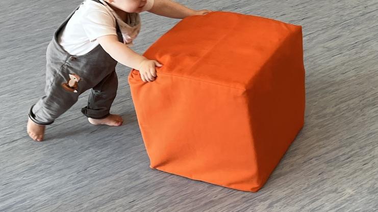 Kleinkind schiebt einen orangefarbenen Würfel auf einem hellen Boden.