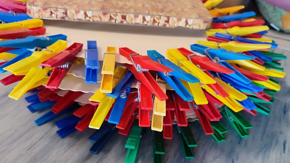 Bunte Wäscheklammern in verschiedenen Farben umringen eine flache, rechteckige Korkplatte auf einer Holzoberfläche.