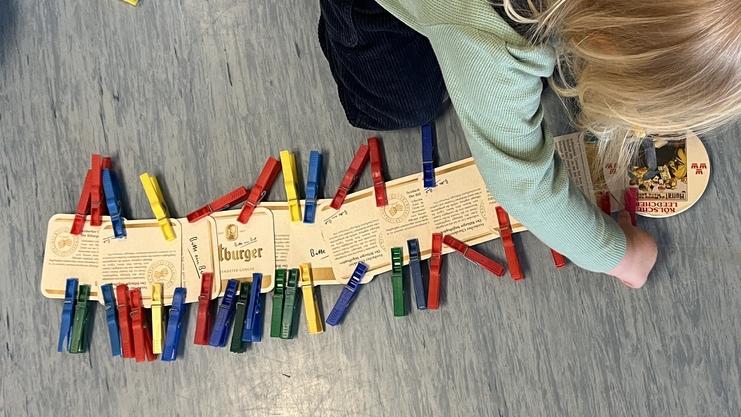 Ein Kind legt bunte Wäscherklammern auf Karten auf einem blauen Boden, während weitere Karten verstreut liegen.