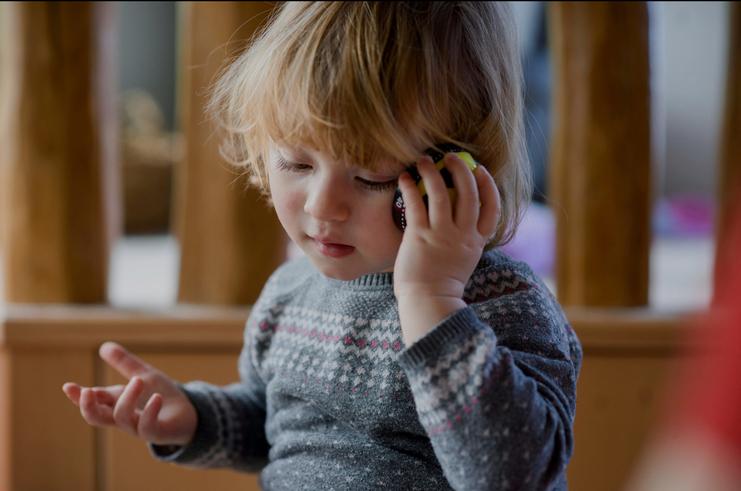 Kleines Kind mit lockigen Haaren hält ein Spielzeugtelefon ans Ohr und schaut nachdenklich. Holzinterieur im Hintergrund.