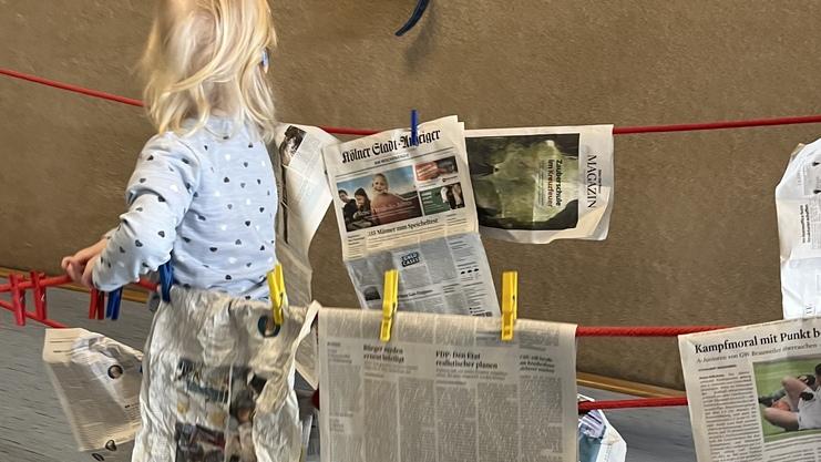 Ein Kind mit blonden Haaren trägt eine Zeitungs-„Schürze“ und spielt mit Wäscheklammern an einer Schnur.