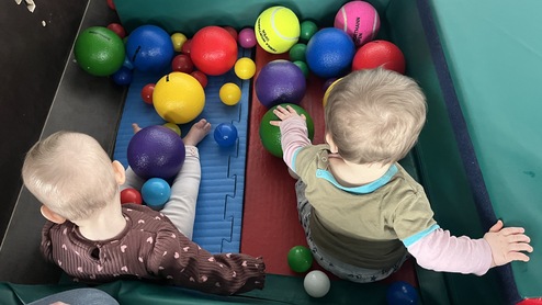 Zwei Kleinkinder sitzen in einer farbigen Ballschleuse mit verschiedenen Bällen auf einem weichen Untergrund.