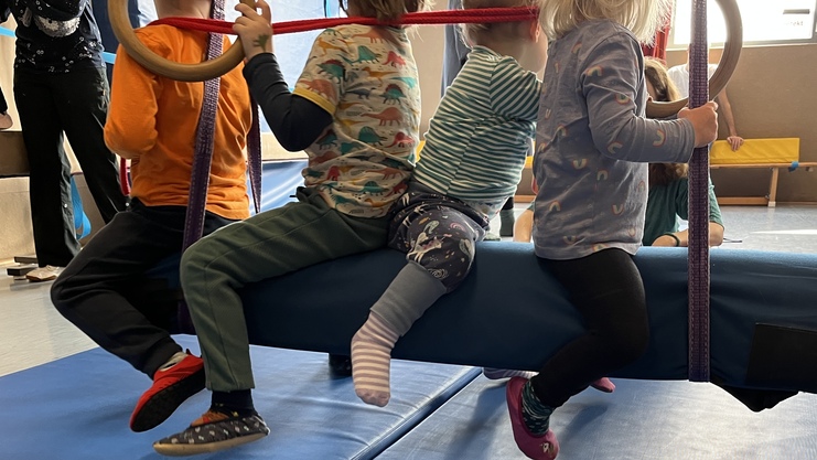 Vier Kinder sitzen auf einem blauen Gymnastikzylinder und halten sich an Ringen und Seilen fest.