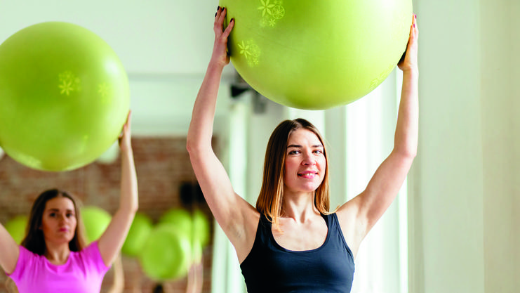 Zwei Frauen in einem Fitnessraum heben große grüne Gymnastikbälle über ihren Köpfen.
