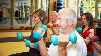 Vier Personen sitzen in einem Fitnessstudio und heben Hanteln in verschiedenen Farben während des Trainings.