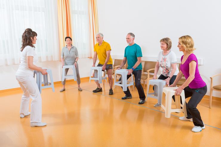 Eine Trainerin demonstriert eine Übung mit sechs Teilnehmern, die Hocker in einer hellen Trainingsumgebung halten.