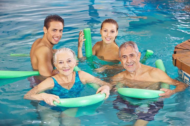 Vier Personen stehen in einem Pool, halten Noodle-Schwimmhilfen und lächeln freundlich in die Kamera.