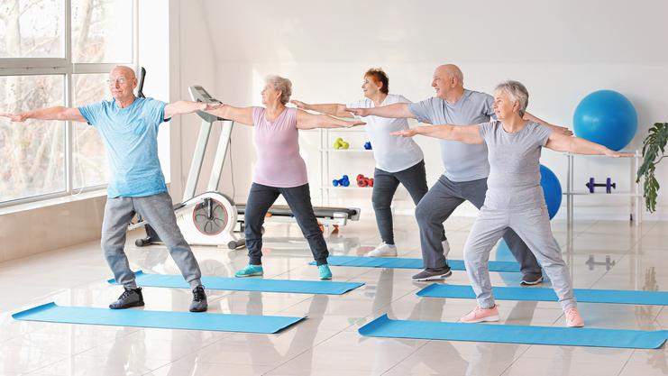 Gruppe älterer Menschen macht Dehnübungen auf blauen Yogamatten in einem hellen Fitnessraum.