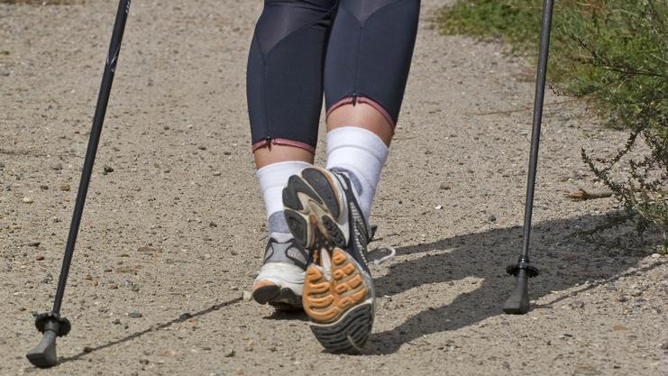 Beine einer Person in Sportkleidung, die mit Walkingstöcken auf einem Kiesweg läuft.
