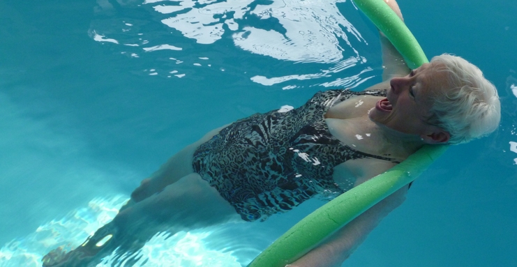 Frau mit kurzem, blondem Haar hält sich mit einem Schwimmnudel in einem blauen Schwimmbecken über Wasser.