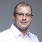 Porträt eines Mannes mit Brille, grauen Haaren und weißem Hemd, vor einem grauen Hintergrund.