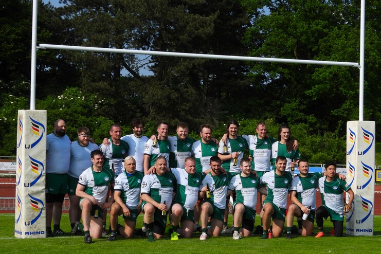 Gruppenfoto von Rugby-Spielern in grünen Trikots, die fröhlich auf einem Platz um die Torpfosten posieren.
