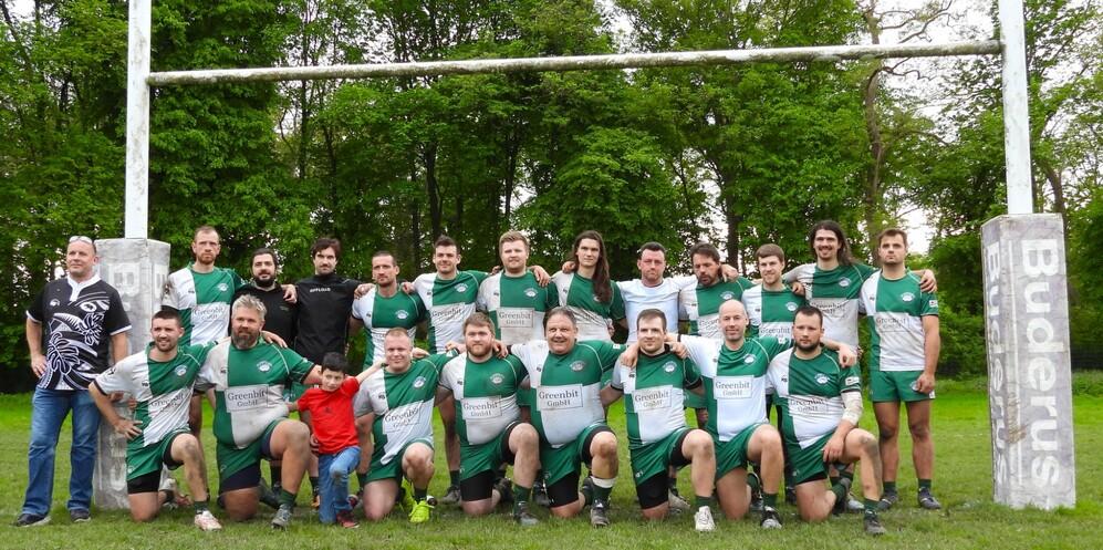 Rugby-Mannschaft in grünen Trikots posiert vor Toren auf einem Platz, mit einem Kind in der Mitte der Gruppe.