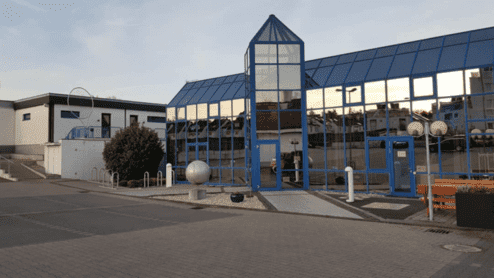 Moderne Glasfassade eines Gebäudes mit blauen Akzenten und runden Sitzgelegenheiten im Vordergrund.