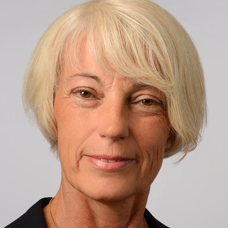 Porträt einer Frau mit kurzen, blonden Haaren und goldener Halskette, bekleidet mit einem schwarzen Blazer vor grauem Hintergrund.