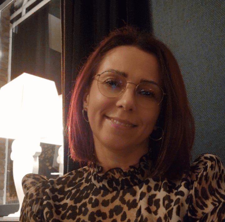 Porträt einer Frau mit rotem Haar, Brille und Leopardenmusterbluse, lächelt vor einem beleuchteten Fenster.