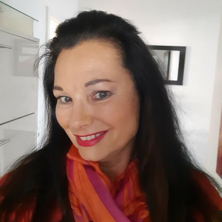 Porträt einer Frau mit langen, dunklen Haaren, rotem Lippenstift und buntem Schal in einer hellen Innenumgebung.