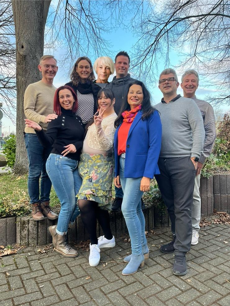 Gruppe von neun Personen posiert lächelnd draußen vor einem blauen Himmel, umgeben von Bäumen und grüner Vegetation.