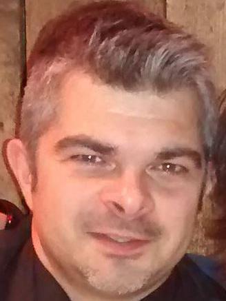 Nahaufnahme eines Mannes mit grauen Haaren und kurzem Bart, lächelt vor einer rustikalen Holzwand.