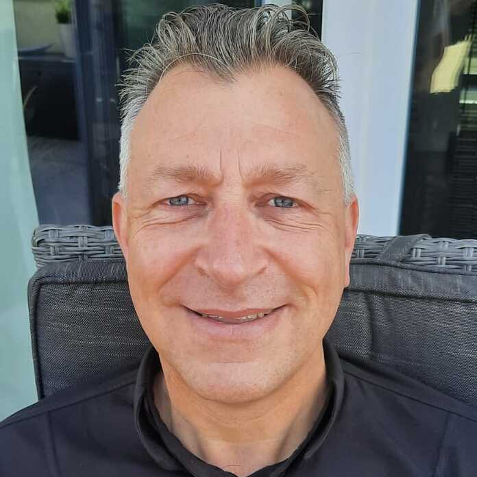 Porträt eines Mannes in schwarzem Hemd, lächelnd, sitzt auf einem Stuhl vor einer Glaswand mit Blick ins Freie.