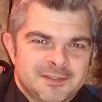 Nahaufnahme eines Mannes mit grauen Haaren und kurzem Bart, lächelt vor einer rustikalen Holzwand.