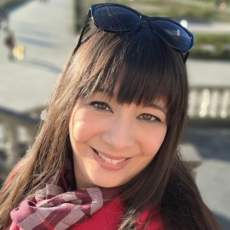 Frau mit langem, braunem Haar in rotem Mantel und Schal lächelt vor einem Park mit Gehwegen und Bäumen.