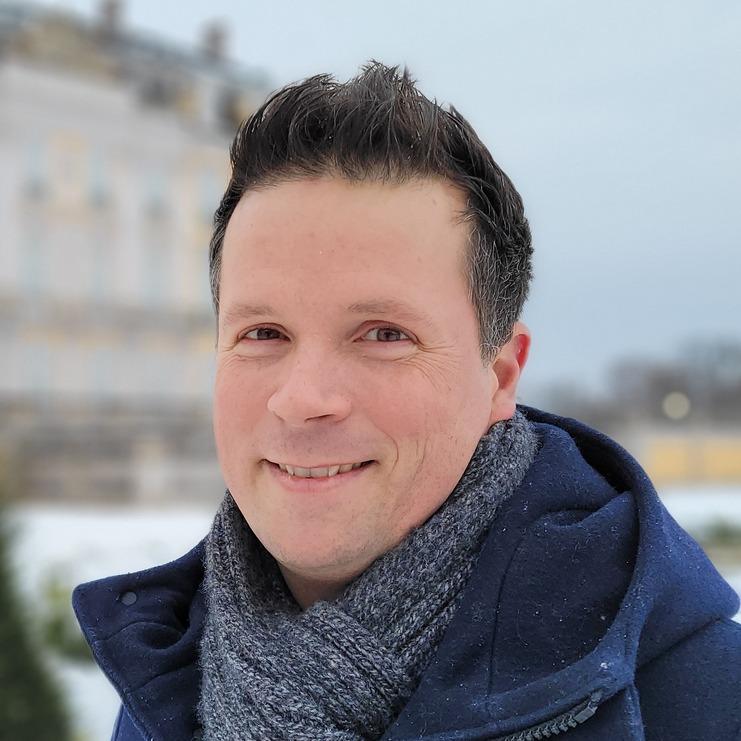 Mann mit dunklem Mantel und Schal lächelt im Schnee vor einem gelben Gebäude mit Fenstern.