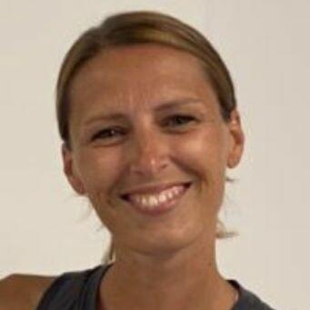 Lächelnde Frau mit hellbraunen Haaren und grauem Tanktop vor neutralem, hellen Hintergrund.