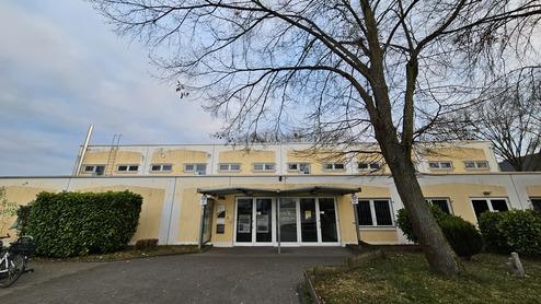 Eingang eines gelben Gebäudes mit Glasdoor, umgeben von Bäumen und Büschen, bei bewölktem Himmel.