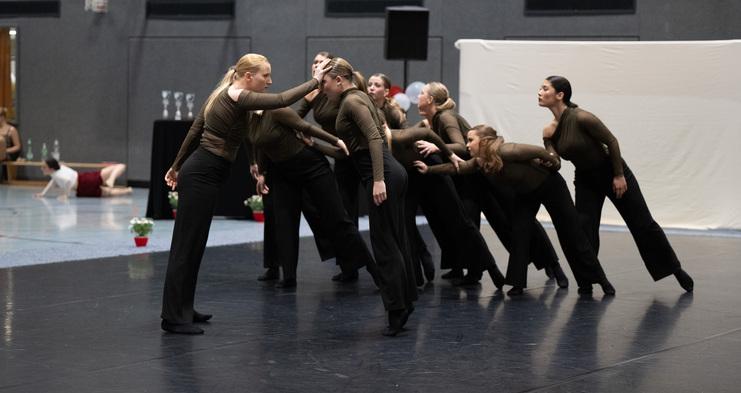 Tanzgruppe in braunen Oberteilen und schwarzen Hosen in dynamischer Formation auf einer Bühne im Sporthallenraum.