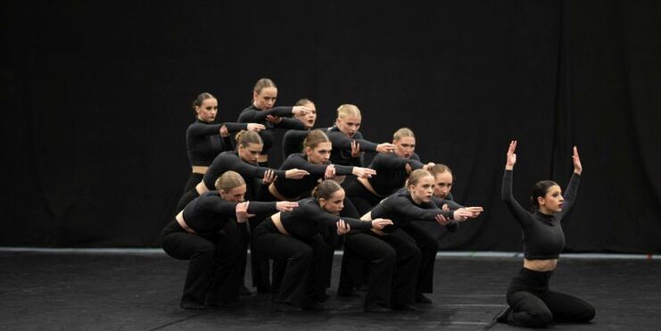 Eine Gruppe von Tänzerinnen in schwarzer Kleidung zeigt synchronisierte Bewegungen auf einer dunklen Bühne.