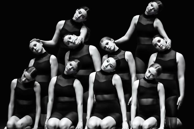 Elf Tänzerinnen in Schwarz posieren in einem gestaffelten Arrangement vor schwarzer Kulisse. Dramatisches Licht.