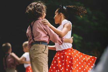Zwei Kinder tanzen auf einer Bühne, das Mädchen trägt einen roten Rock mit weißen Punkten, der Junge einen karierten Hemd.