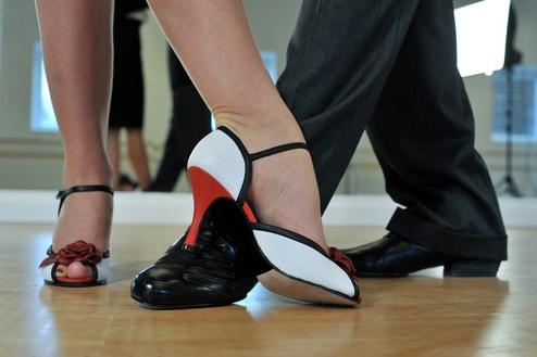 Tanzpartner stehen sich gegenüber, ihre Füße auf einem Holzboden, elegante Tanzschuhe in Schwarz und Weiß mit roten Akzenten.
