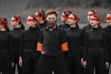 Gruppe von Tänzern in Schwarz mit roten Augenbinden und Blumenhaarschmuck, im Vordergrund ein Tänzer in auffälligem Outfit.