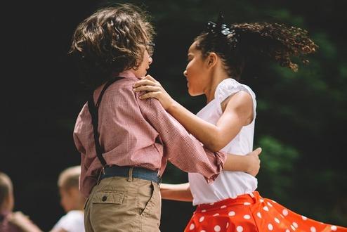 Zwei Kinder tanzen auf einer Bühne, das Mädchen trägt einen roten Rock mit weißen Punkten, der Junge einen karierten Hemd.