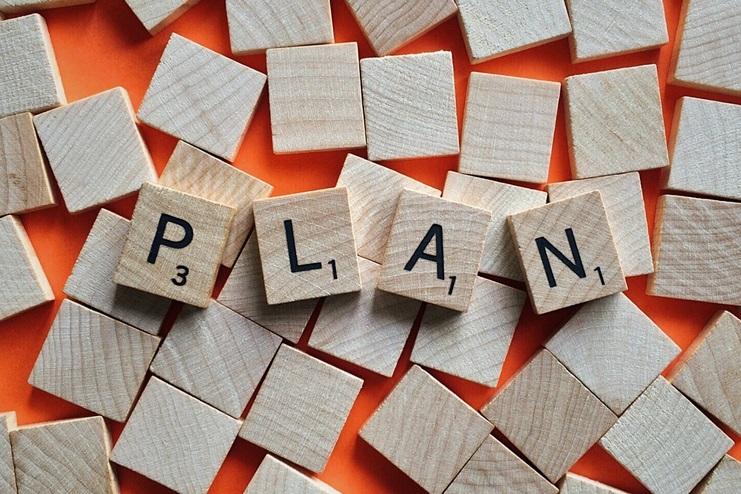 Holzplättchen mit Buchstaben formen das Wort „PLAN“ auf einem orangefarbenen Hintergrund.