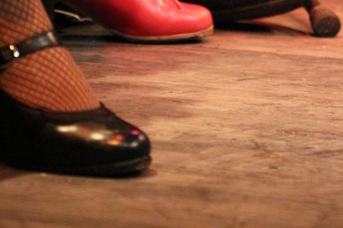 Nahaufnahme von verschiedenen Schuhen auf einem Holzboden, darunter rote Tanzschuhe und schwarze Lackschuhe.