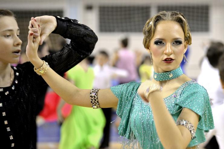 Tänzerin in glitzerndem Outfit hält die Hand eines Partners auf einer Tanzfläche mit weiteren Tänzern im Hintergrund.
