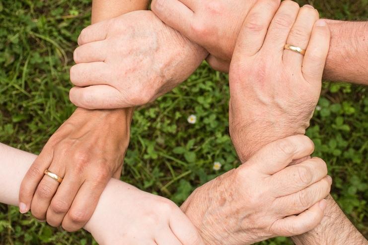 Sechs Hände unterschiedlicher Hauttöne greifen ineinander auf einer Wiese.