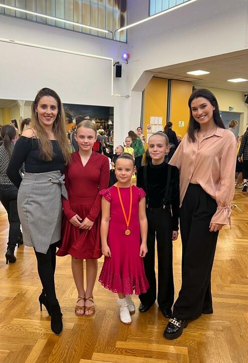 Fünf Personen stehen lächelnd in einem hellen Raum, bekleidet in festlicher Kleidung. Ein Mädchen trägt eine Medaille.