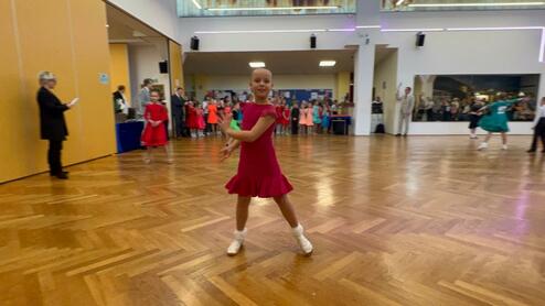 Mädchen in rotem Kleid tanzt auf einer Holzfläche, umgeben von anderen Tänzern und Publikum im Hintergrund.