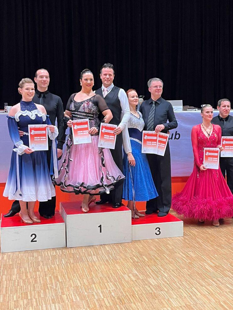 Siegerpaar und Finalisten in festlicher Kleidung halten Urkunden auf dem Treppchen einer Tanzveranstaltung.
